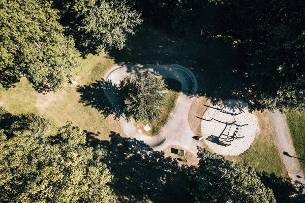 Luftaufnahme von einem Kinder Pumptrack neben einem Spielplatz in einem Park mit vielen Bäumen. 