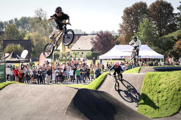 Fahrradfahrer*innen springen und machen Tricks auf einem Pumptrack. Kinder schauen begeistert zu.  