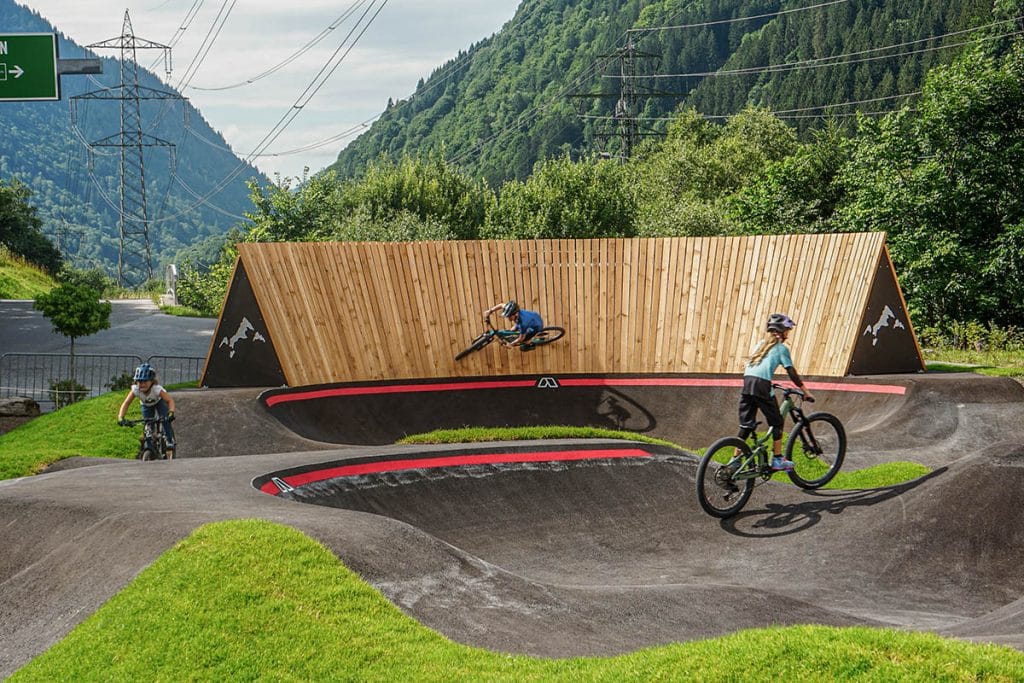 Zu sehen ist ein Holzwallride auf einem asphaltierten Pumptrack. Drei Fahrerinnen sind auf dem Pumtrack unterwegs, einer davon am Wallride. 