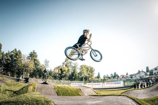 Ein BMX Fahrer bei einem Sprung in der Luft der einen Trick macht. Der Sprung ist auf einer Jumpline, diese wiederum ein Teil einer Pumptrackanlage. 